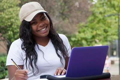 A smiling young female student wearing a tan baseball cap sits outdoors at a table with a purple laptop and a pencil, with trees and a campus walkway visible in the background.