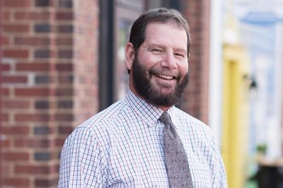 A smiling middle-aged man with a beard wearing a plaid button-down shirt and tie stands outdoors in front of a brick building.