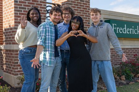 diverse students in front of brick sign. one girl waving and one making heart with hands