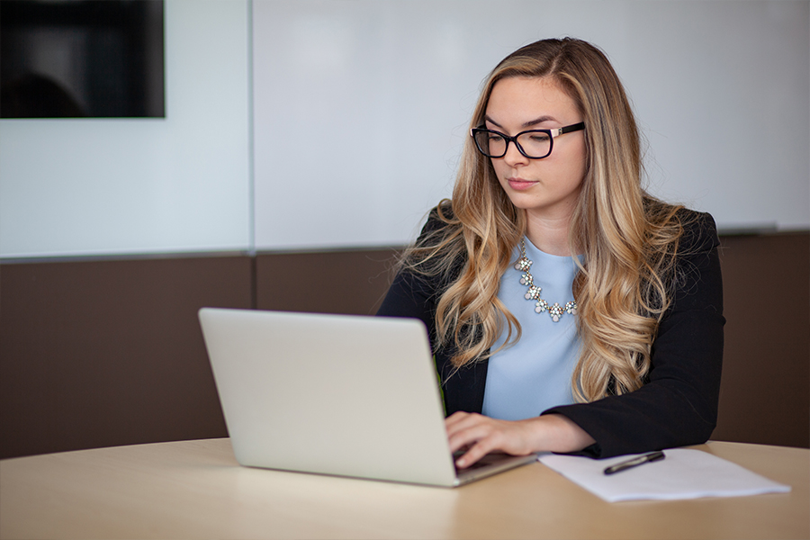 A professional woman with blonde hair and glasses works on a laptop at a desk with a notebook and pen.