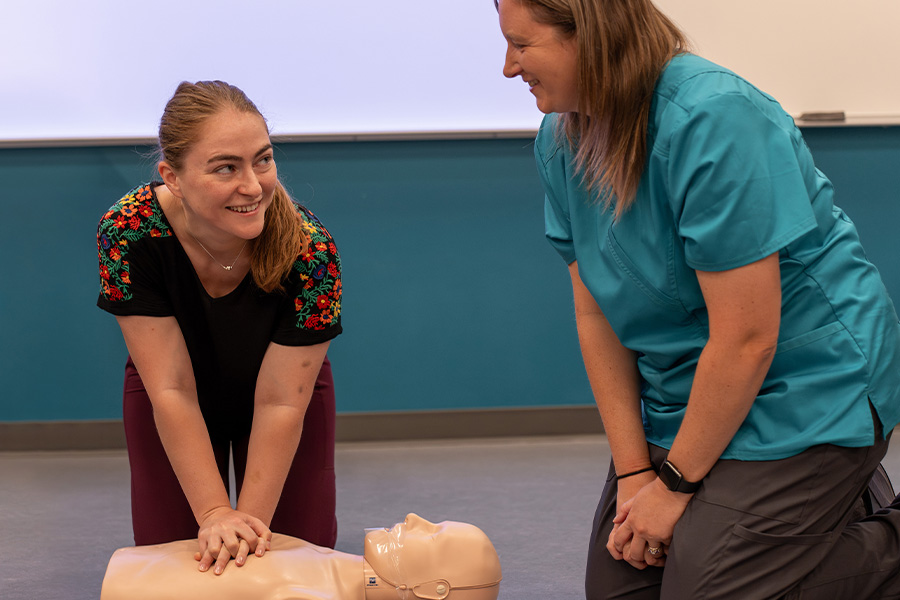 Student practicing chest compressions on a CPR manikin while an instructor in scrubs looks on