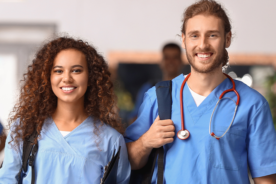 Two smiling healthcare students in blue scrubs carrying backpacks and walking through a hallway, one wearing a stethoscope.
