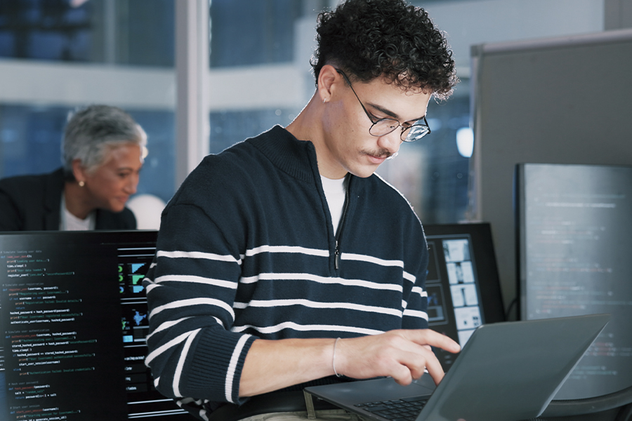 Student working on a laptop surrounded by screens displaying code in a cybersecurity lab