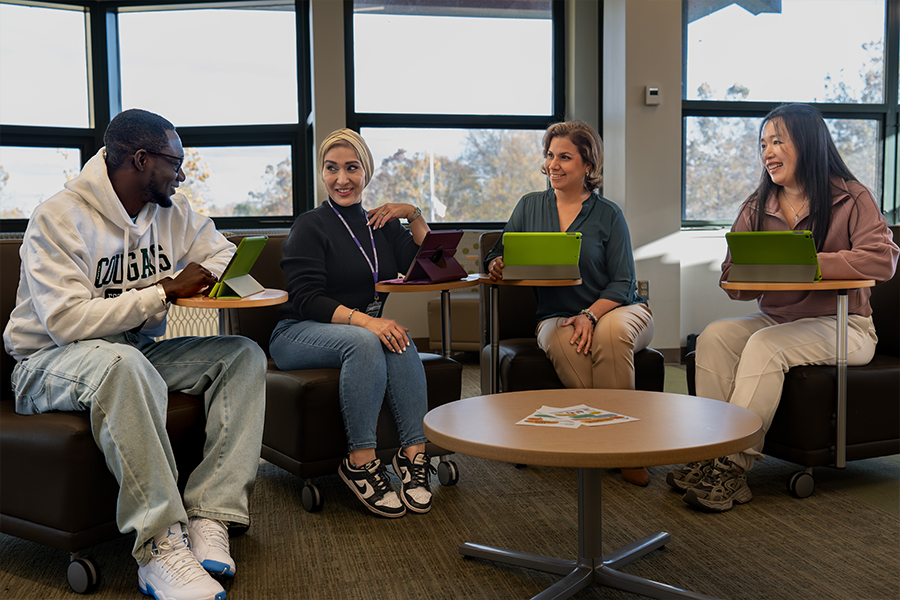 Four people sit in a semi-circle on mobile lounge chairs in a sunlit room, each using a tablet mounted on a built-in swivel desk. The group, appearing to be students or colleagues of diverse backgrounds, are smiling and engaged in a casual discussion. A round coffee table sits in the center of the group, and large windows in the background look out onto trees.