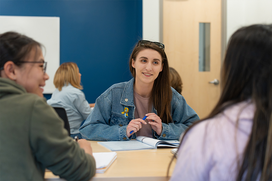 A smiling student in a denim jacket sits at a table with classmates, holding a pen and participating in a group discussion.