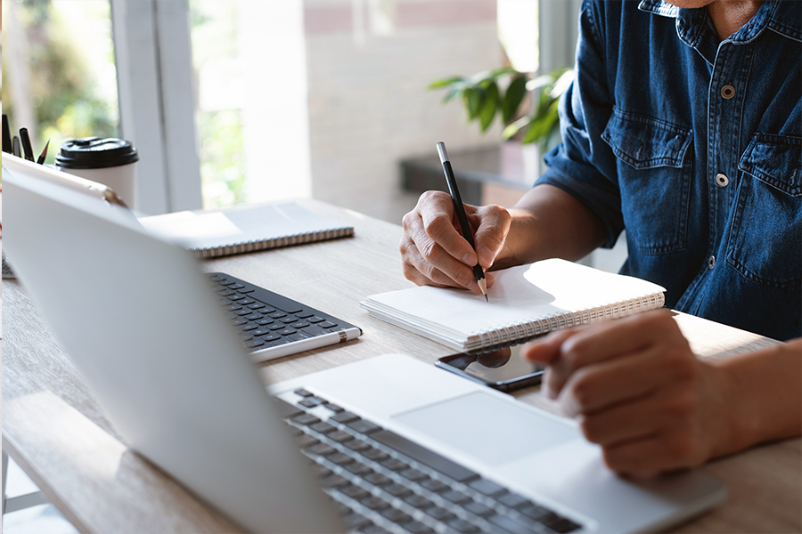 The individual is actively writing with a black pen in a small, white spiral-bound notebook. Their right hand is poised on the paper, while their left hand rests near a laptop in the foreground.