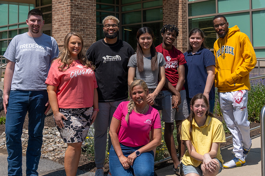 A diverse group of nine smiling students and staff pose outdoors in front of a brick building at Frederick Community College, some wearing college-branded apparel.