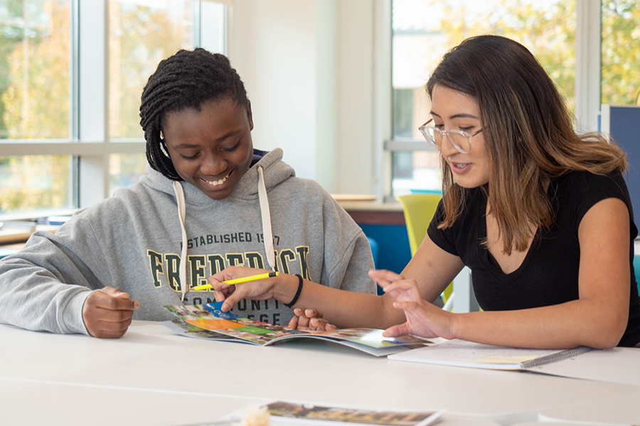 Two students review a printed publication together at a table in a bright FCC classroom, both smiling and engaged in conversation.
