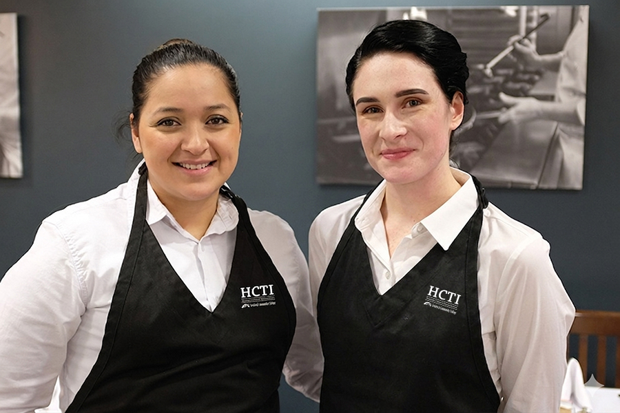 Two FCC hospitality students in white shirts and HCTI aprons smile in a restaurant dining room.