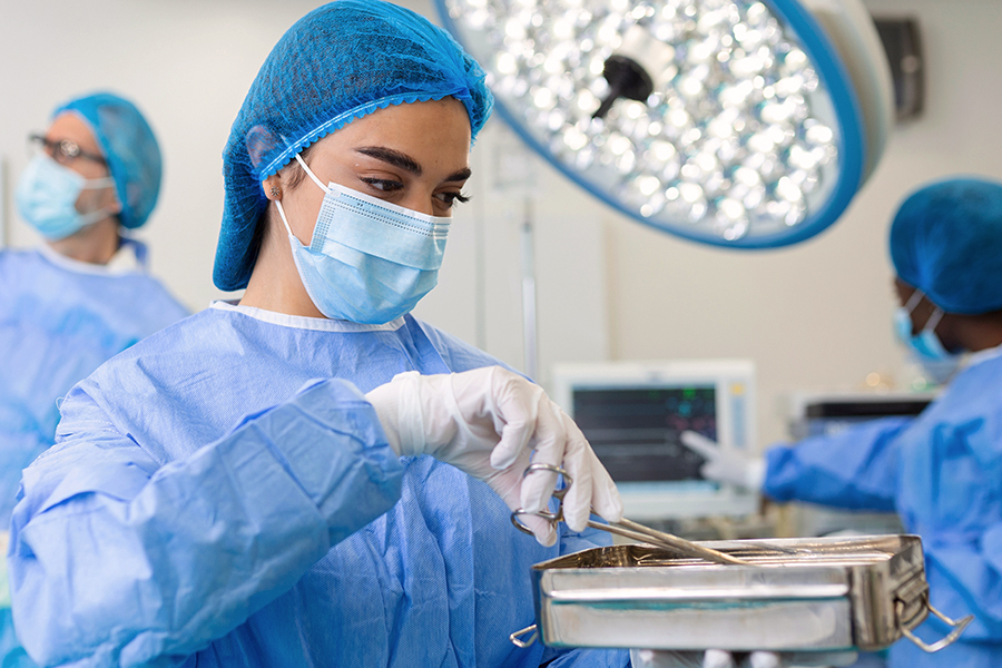 A surgical team member in full PPE, including a gown, mask, hair cover, and gloves, handles sterile instruments in a tray in an operating room. Other team members work in the background.