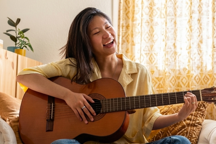 A joyful young woman plays an acoustic guitar and sings while sitting on a comfortable sofa.