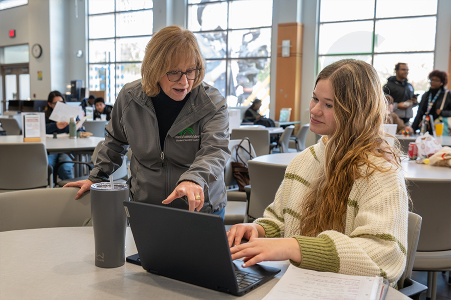 An older woman, possibly a teacher or advisor, leans over a laptop to point something out on the screen to a younger woman. Both are engaged in the digital content, showing a clear moment of guidance and learning.