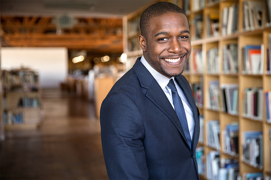 A medium shot of a smiling Black man in a navy suit and tie, standing in a bright, modern library or office with wooden bookshelves in the background.