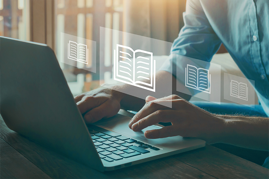 A person types on a laptop with translucent digital book icons floating above the keyboard. This conceptual image represents digital learning or an online library.