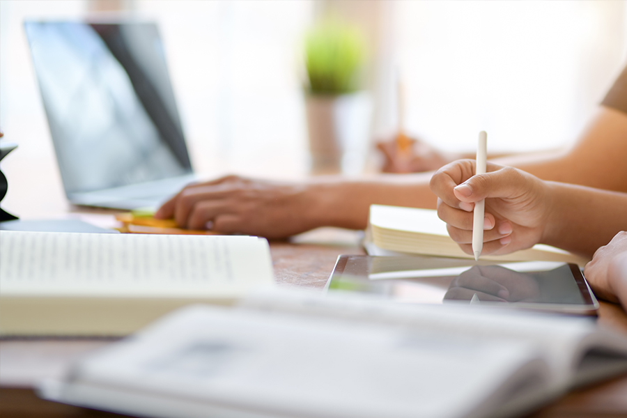 A person uses a white stylus to write on a tablet in a bright workspace surrounded by open books and a laptop.