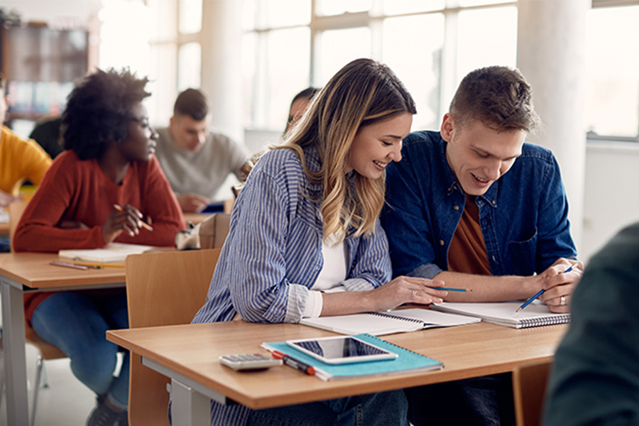 Two students sit together at desks in a sunlit classroom, smiling as they review notes on a tablet.