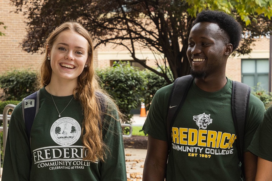 Two students, a woman with long blonde hair and a man with short dark hair, wearing green Frederick Community College t-shirts and backpacks, smiling and walking together on campus.