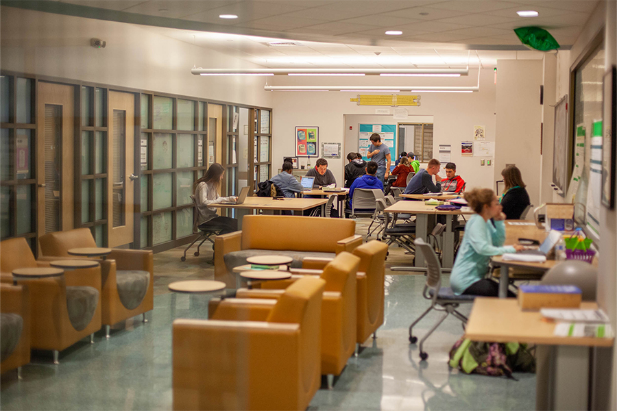A vibrant, high-angle view of the STEM Learning Center through a glass partition. In the foreground, several tan, modern armchairs with built-in circular side tables are arranged on a polished floor. Beyond them, students are seated at light-wood rectangular tables, working on laptops and collaborating in small groups. The space is well-lit with overhead linear lighting and features glass-walled study rooms along the left side and informational posters on the far wall.