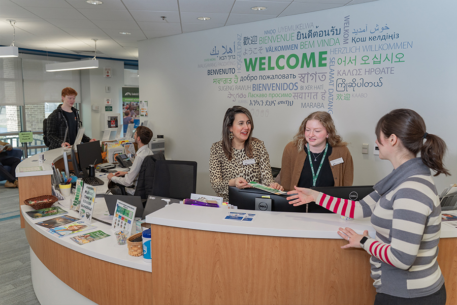 At a curved wooden reception desk, two staff members stand under a wall featuring the word "Welcome" in multiple languages while interacting with a visitor.