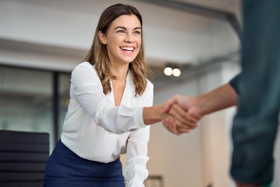 A smiling professional woman in a white blouse and navy skirt shaking hands with an unseen colleague in a modern, bright office setting.