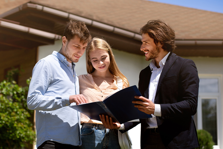 A smiling real estate agent showing documents to a young couple holding a folder in front of a new house.