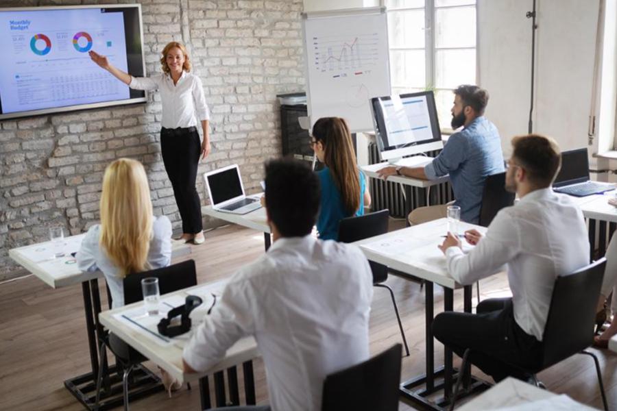 A female business professional presenting a monthly budget report to a team in a modern office classroom.
