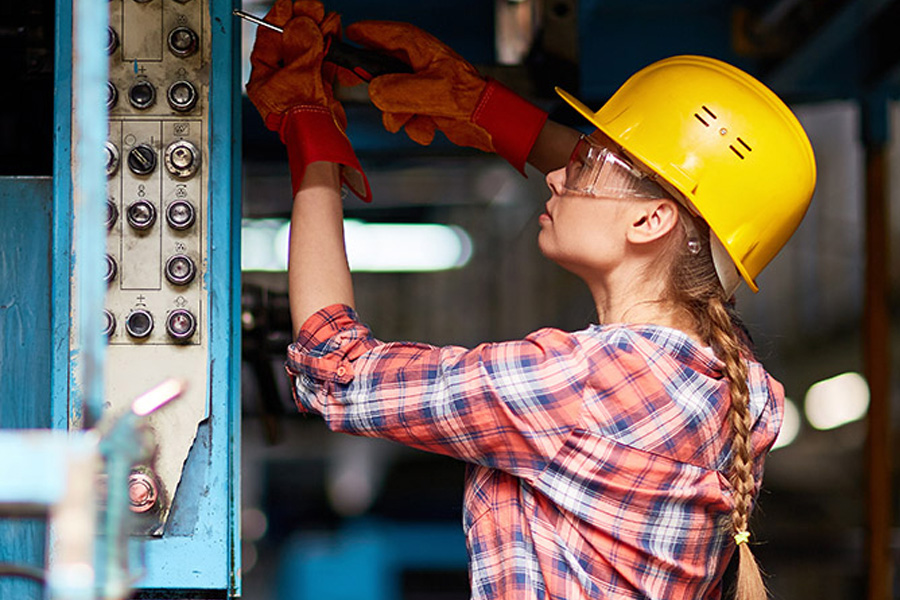 A female electrician wearing a yellow hard hat and safety glasses working on a blue industrial electrical panel.