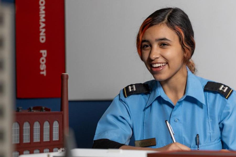 A smiling young woman in a blue security or cadet uniform sitting at a desk and holding a pen.
