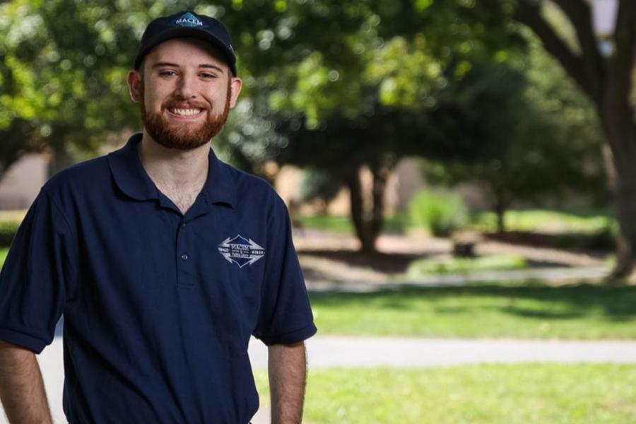 A smiling man in a navy blue polo shirt and matching baseball cap standing outdoors on a campus.