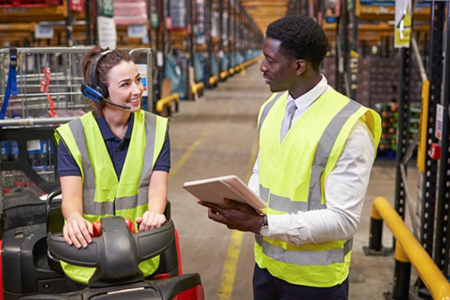 A logistics trainee in a high-visibility vest and headset operates a motorized pallet jack while discussing inventory with a supervisor holding a tablet in a large warehouse.