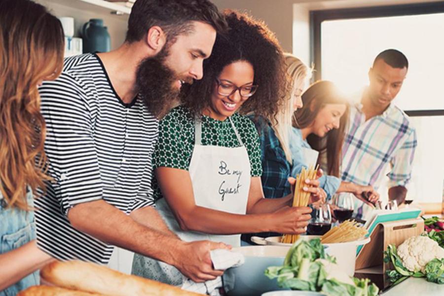 A diverse group of adult students enjoys a hands-on community cooking class, with one woman in a Be My Guest apron preparing pasta while others look on.