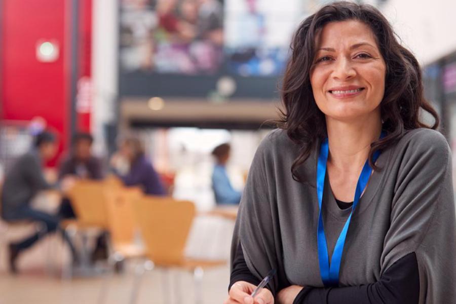 A smiling woman with dark hair wearing a gray cardigan and a blue lanyard stands with her arms crossed in a busy academic common area, with students visible in the blurred background.