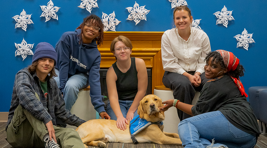 Five smiling young adults posing with a yellow Labrador Retriever wearing a blue "Canine Companions" vest, gathered in front of a blue wall decorated with large paper snowflakes.